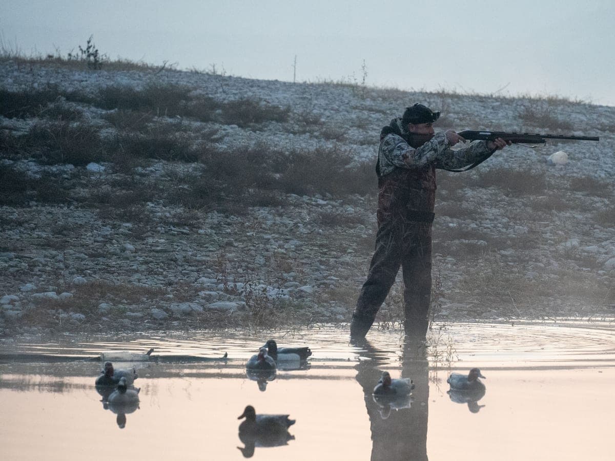 A waterfowl hunter aiming over a decoy spread on still water — Decoy & Drake, waterfowl hunting gear