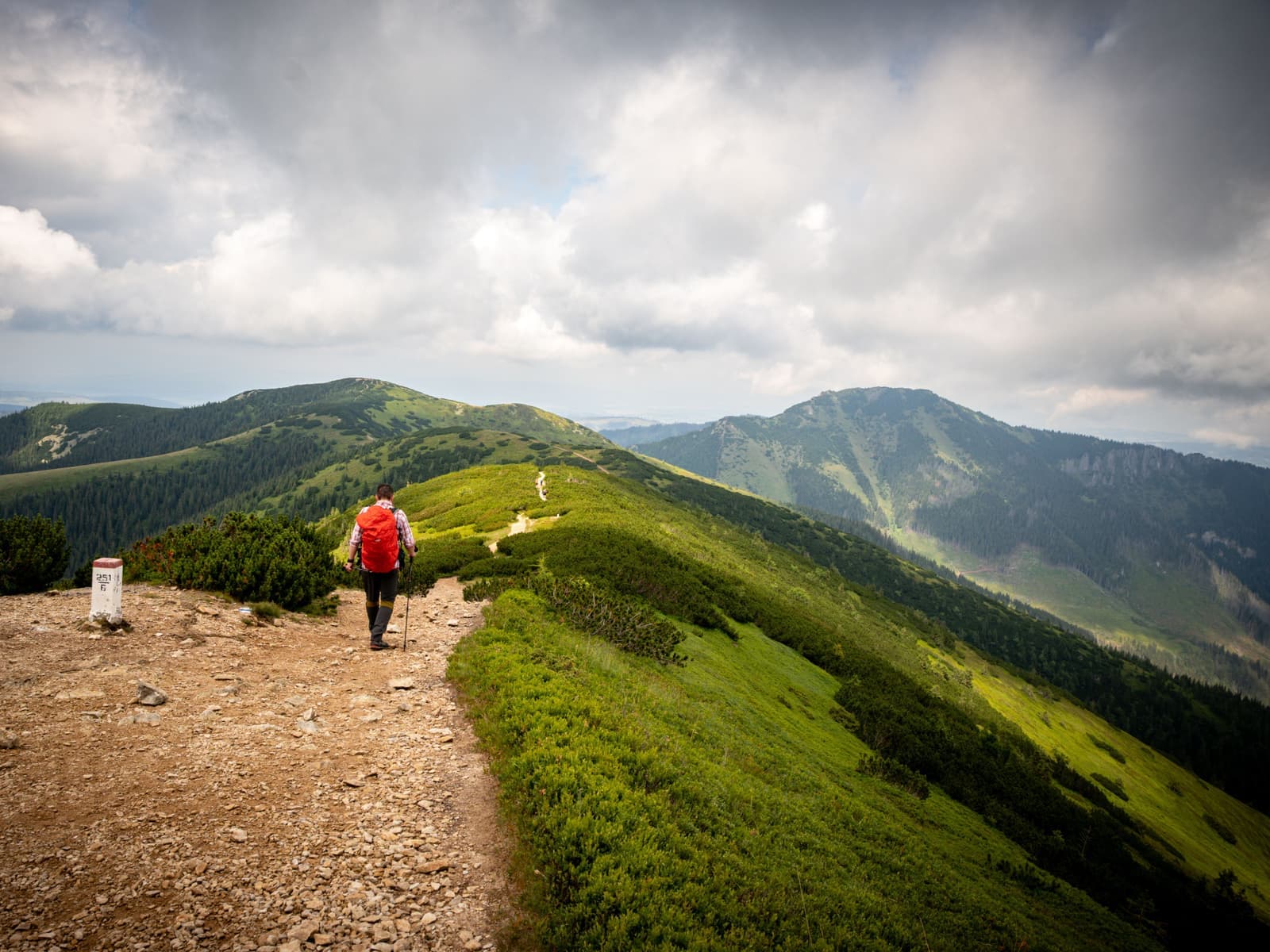 A hiker with a red pack and trekking pole walking a rocky ridge trail under a clouded sky